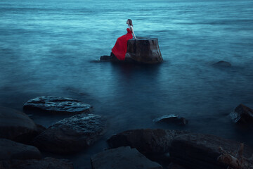 A sad woman in red dress near the sea in evening. Long time exposure