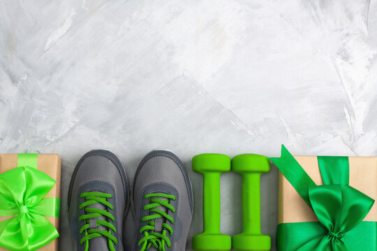Holiday christmas birthday party sport flat lay composition with gray shoes, green dumbbells  and craft gifts with green bow on gray concrete background. Top view, horizontal orientation