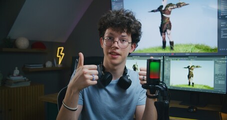 Young 3D designer speaks on camera using microphone, records video tutorial for blog about video game design and 3D modeling at home studio. Software interface on computer and big digital screen.