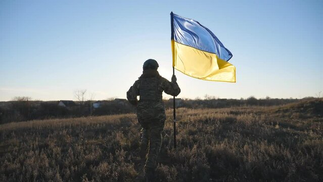 Young woman in camouflage uniform jogs with waving flag of Ukraine against sunset. Female soldier of ukrainian army runs with lifted national banner on the field. Invasion resistance. Rear view