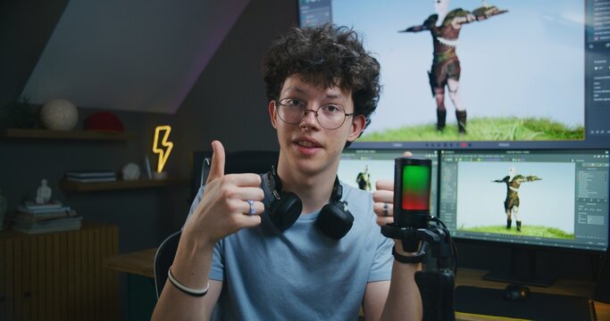 Young 3D designer speaks on camera using microphone, records video tutorial for blog about video game design and 3D modeling at home studio. Software interface on computer and big digital screen.