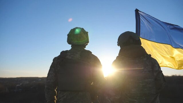 Dolly shot of young woman and man in military uniform lifted flag of Ukraine at countryside. Female and male soldier of ukrainian army stands at the peak of hill with a waving blue-yellow banner