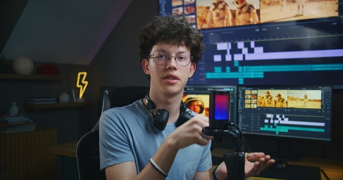Young editor, video maker speaks on camera using microphone, records video training about film and video editing for blog. Movie footage and software interface on computer and big digital screen.