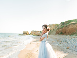 Beautiful brunette bride in light chiffon wedding dress embroidered with beads posing near the sea....