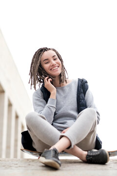 Attractive and smiling teenager girl relaxing with a skateboard and sitting down, talking to somebody on phone, outdoors.