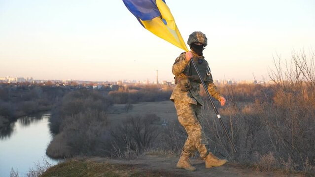Ukrainian army man dancing funny with blue-yellow flag on hill at countryside. Happy male soldier in camouflage uniform celebrating victory of war between Ukraine and Russia. End of warfare in Europe