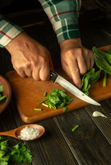 The chef cuts fresh green spinach on a cutting board with a knife. Close-up of the chef's hands while working on the kitchen table. The idea of a vegetarian or vegetable diet.