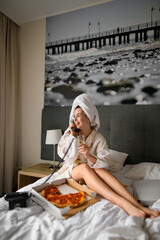 Young woman in bathrobe sitting on bed with champagne glass in hand and calling by phone