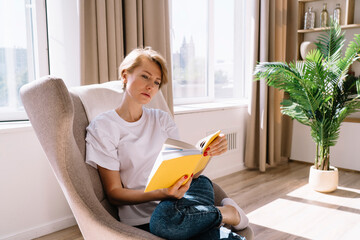 Female frowning while reading book in armchair in light room