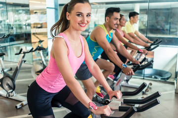 Side view of a beautiful fit young woman smiling while pedaling during cardio workout at indoor...