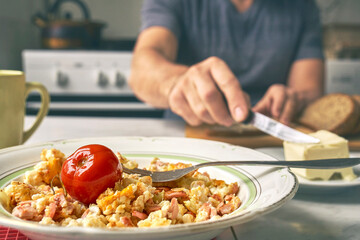 Freshly made omelette with sausages in a plate on the foreground and a man smearing a butter on bread on blurred background