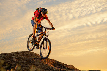 Fototapeta premium Cyclist in Red T-Shirt Riding the Bike Down the Rock on the Sunset Sky Background. Extreme Sport and Enduro Biking Concept.