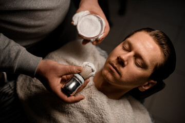 Fototapeta premium Barber applying shaving foam onto client's face using old-fashioned shaving brush in barbershop