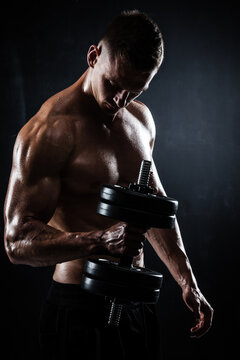 Athletic shirtless young male fitness model makes exercises with dumbbells on dark background