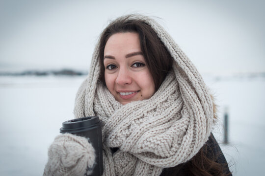 Young woman in warm and soft scarf with hot coffee in cup for take away staying at clear field on the winter background. Cold day. White and soft colors.