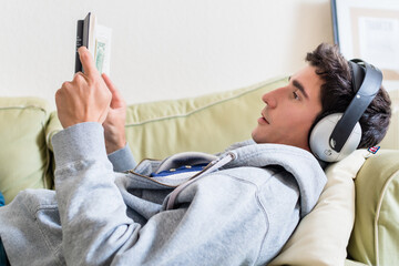 Relaxed young man lying down on the couch while listening to music and reading a book at home