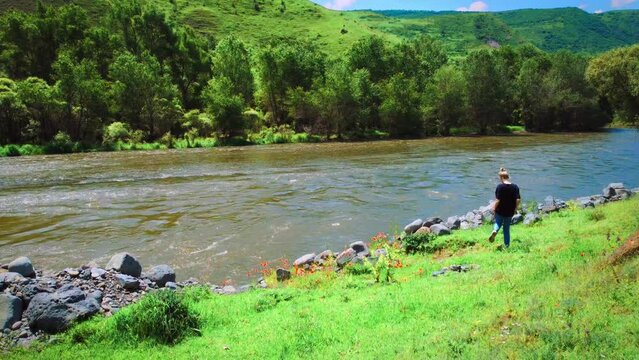 Young woman in jeans and a T-shirt stands near a mountain river against the background of mountains.Beautiful landscape with a river, trees,poppies and a girl from the back.Concept of travel,lifestyle