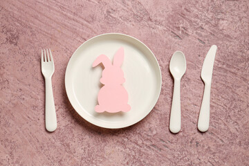Plate with eating utensils for baby and paper rabbit on grunge pink background