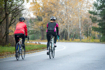 Two Young Female Cyclists Riding Road Bicycles in the Park in the Cold Autumn Morning. Healthy Lifestyle Concept.
