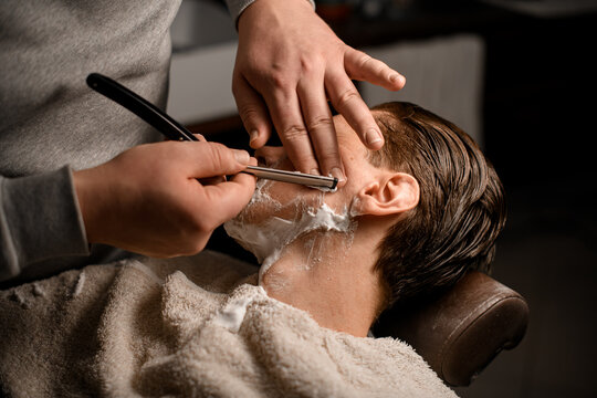 Barber man shaving young attractive client using vintage straight razor with stainless steel in his salon