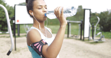 Beautiful ethnic woman in sportswear using smartphone and headphones and drinking water with eyes closed in park.