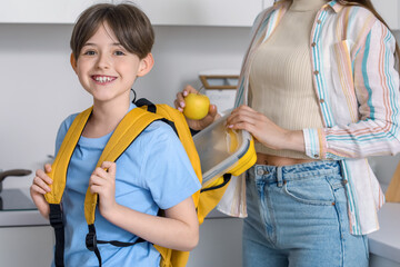 Mother putting school lunch in her little son's backpack in kitchen