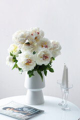 Vase of white peonies with candles and magazine on coffee table near wall