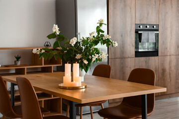 Vase with blooming jasmine flowers and burning candles on wooden table in modern kitchen