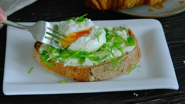 Cutting a poached egg with fork, benedict egg with runny egg yolk and stracciatella cheese on toast, eating healthy breakfast, close-up video, high quality 4k horizontal video