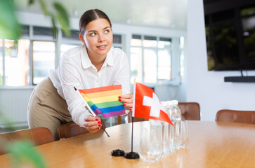Positive young woman putting little LGBT flag on table next to the flag of Switzerland and bottles of water in conference room