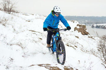 Cyclist in Blue Riding the Mountain Bike on the Rocky Winter Hill Covered with Snow. Extreme Sport and Enduro Biking Concept.