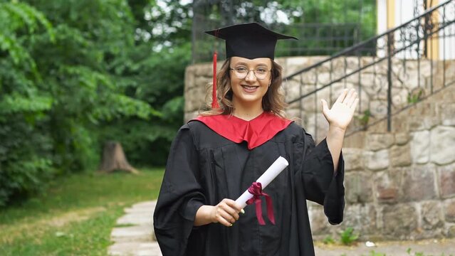 Young woman graduate student in mortarboard and bachelor gown waving hand gesture to say hello in slow motion
