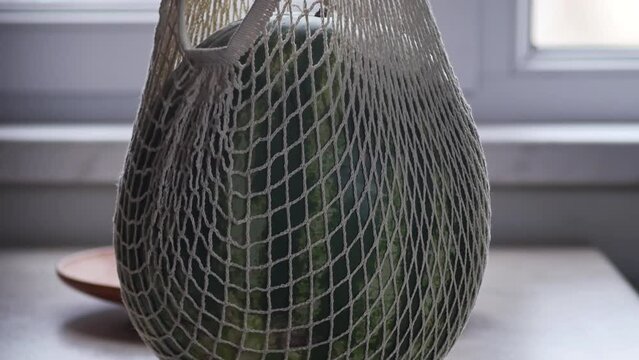 Men's hands put a large watermelon in a string bag on the table and, taking it with both hands, moved a large clay plate. watermelon large size and weight. Eco mesh fucking fruits and vegetables