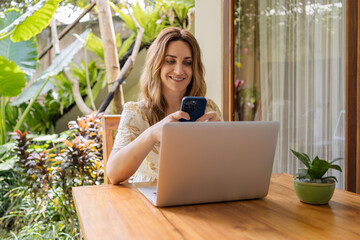 Portrait of young woman freelancer working with laptop on terrace of tropical bungalow with palm trees view in Bali.
