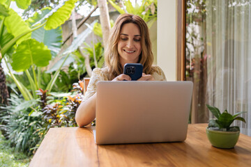 Portrait of young woman freelancer working with laptop on terrace of tropical bungalow with palm trees view in Bali.
