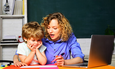 Female elementary school teacher helping little boy with writing lesson in classroom at school. Education and learning concept. Teacher in classroom helps kid in elementary school lessons. Schooling.