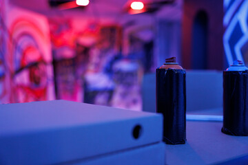 Graffiti spray paint cans placed in ruined warehouse, empty old destroyed building glowing under purple bright neon lights. Bottle of aerosols paint in creepy illuminated warehouse. Close up.