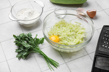 Bowl with grated zucchini and ingredients for preparing fritters on white tile background