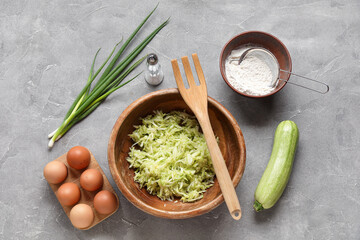 Bowl with grated zucchini and ingredients for preparing fritters on grey background
