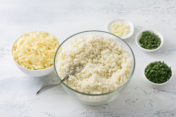 Shredded cauliflower, cauliflower rice or cauliflower couscous surrounded by chopped herbs, garlic and grated cheese on a light blue table. Cooking delicious vegan healthy food