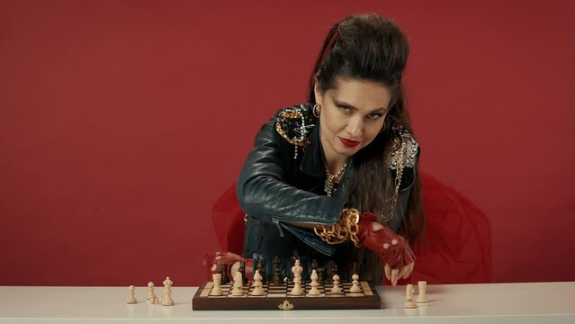 A woman puts chess pieces on a board, gets up from the table, shows with her hand that everything is ready for the game. Portrait of a woman on a red background. Video 2 in 1. Advertising.