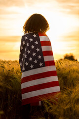Vintage style photograph of mixed race African American girl teenager female young woman in a field of wheat or barley crops wrapped in USA stars and stripes flag in golden sunset evening sunshine