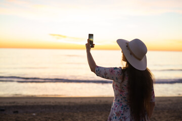 Young woman taking photo the sea and sunset with cell phone or smartphone digital camera for Post...