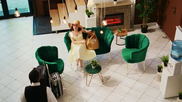 Bellhop offering services to woman in lounge area, waiting to do room check in at seaside hotel. Tourist sitting on couch and talking to hotel concierge, summer clothes. Handheld shot.