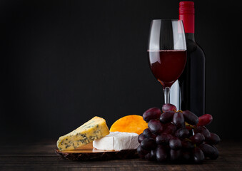 Bottle and glass of red wine with cheese selection with grapes on black background