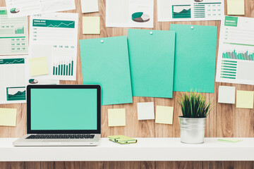 Desktop with laptop and paperwork hanging on the office wooden wall and blank green sheets at center, eco business concept