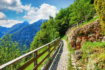 Fototapeta na wymiar Hallstatt, Austria. Picturesque paved stone track along picturesque knolls above old town among green trees and blue mountains covered with forests. Wooden fence. Blue sky with clouds.