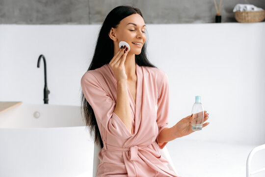 Facial cleansing with tonic. Satisfied lovely caucasian brunette young woman, in a bathrobe, sits in the bathroom, cleans her face after makeup using cotton pads and tinic, smiles with eyes closed