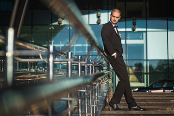handsome elegant groom leaning on metal handrails holding hands in pockets of elegant suit