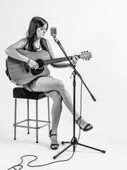 Fototapeta premium Photo of a young woman with long hair sitting on a stool playing an acoustic guitar over white background.